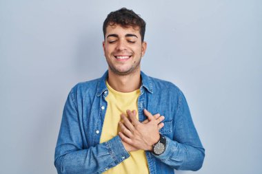 Mavi arka planda duran genç İspanyol adam elleri göğsünde, gözleri kapalı ve yüzünde minnet dolu bir jest ile gülümsüyor. sağlık kavramı. 