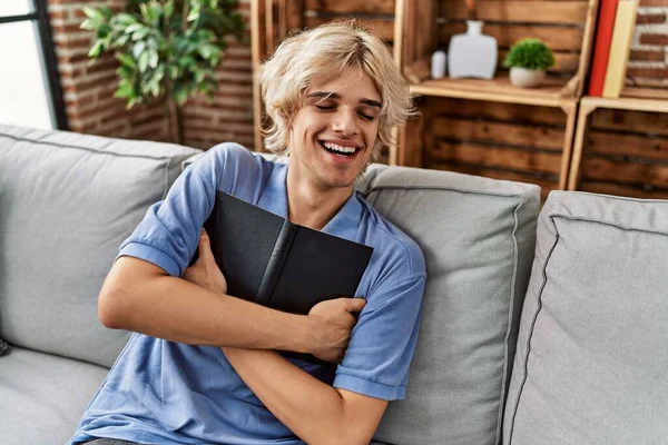 Young blond man hugging book sitting on sofa at home