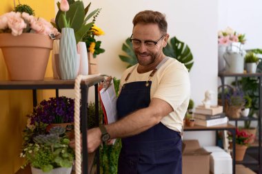 Middle age man florist touching plant holding clipboard at flower shop