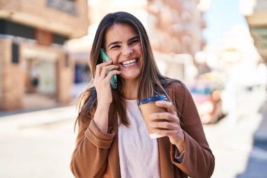 Young beautiful hispanic woman talking on smartphone drinking coffee at street