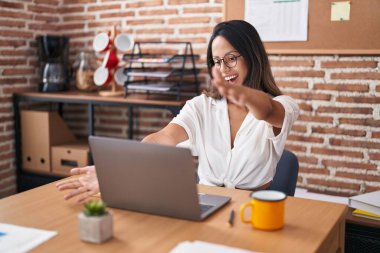 Ofiste çalışan, gözlüklü, kucaklaşmak için kollarını açmış kameraya bakan Latin asıllı genç bir kadın. Mutluluğu kucaklayan neşeli bir ifade.. 