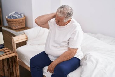 Orta yaşlı, kır saçlı, boynu incinmiş bir adam yatak odasında oturuyor.