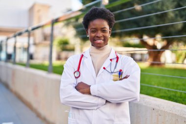 Doktor üniforması giyen Afrikalı Amerikalı kadın caddede kollarını kavuşturmuş duruyor.