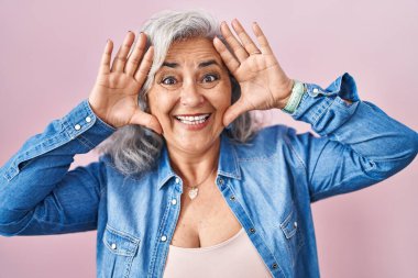 Middle age woman with grey hair standing over pink background smiling cheerful playing peek a boo with hands showing face. surprised and exited 