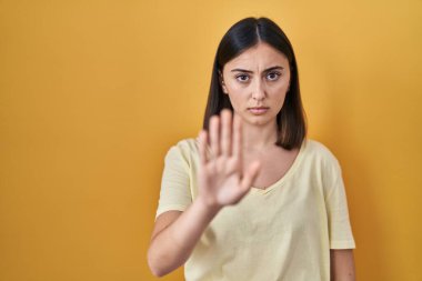 Sarı arka plan üzerine günlük tişört giyen İspanyol kız avuç içiyle şarkı söylemeyi kesiyor. Yüzünde olumsuz ve ciddi bir jest olan uyarı ifadesi. 