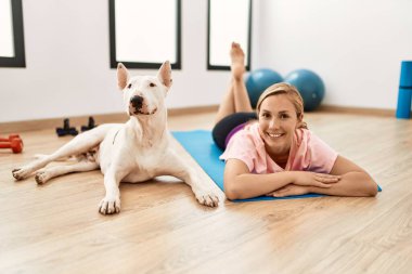 Beyazlı genç bir kadın spor merkezinde yoga minderine uzanmış kendinden emin bir şekilde gülümsüyor.