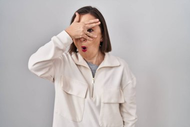 Middle age hispanic woman standing over isolated background peeking in shock covering face and eyes with hand, looking through fingers with embarrassed expression. 