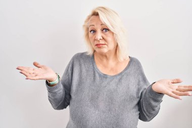 Middle age caucasian woman standing over white background clueless and confused expression with arms and hands raised. doubt concept. 