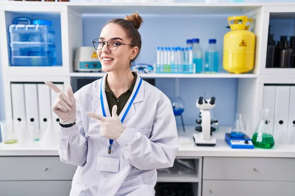Young hispanic girl working at scientist laboratory smiling and looking at the camera pointing with two hands and fingers to the side.