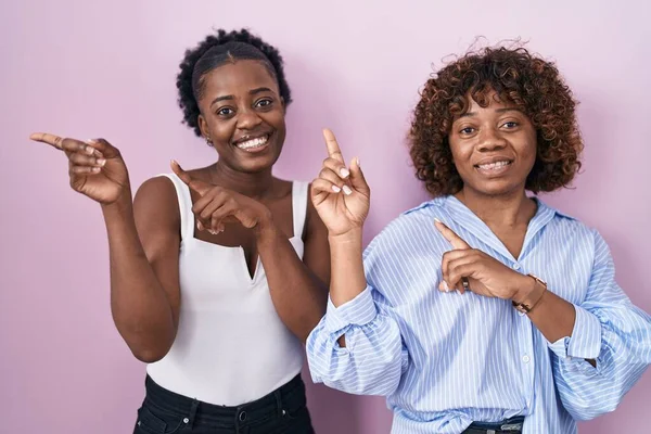 两个非洲女人笑着站在粉色的背景上 看着摄像机 用双手和手指指向旁边 — 图库照片