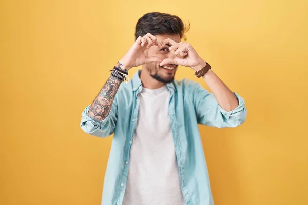 Young Hispanic Man Tattoos Standing Yellow Background Doing Heart Shape — Φωτογραφία Αρχείου