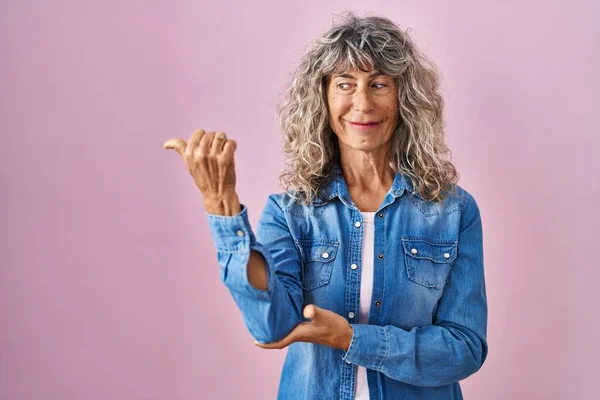 Middle age woman standing over pink background smiling with happy face looking and pointing to the side with thumb up.