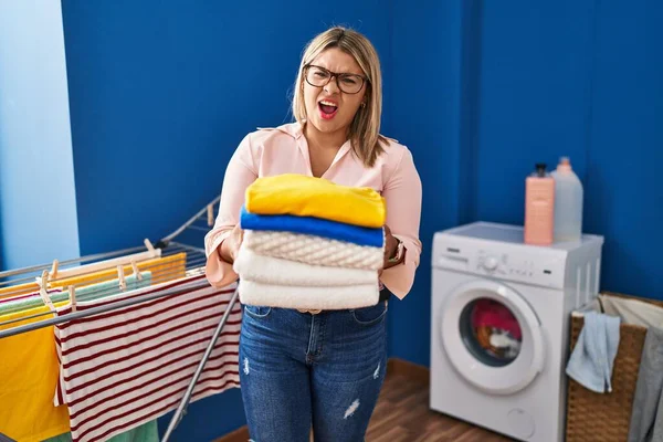 Young hispanic woman holding folded laundry after ironing angry and mad screaming frustrated and furious, shouting with anger. rage and aggressive concept.