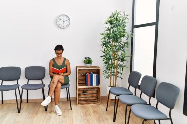 Genç İspanyol kadın bekleme odasında sandalyeye oturmuş kitap okuyor.