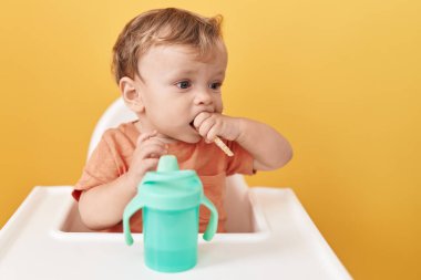 Sevimli sarışın bebek sandalyeye oturmuş izole edilmiş sarı arka planda atıştırıyor.