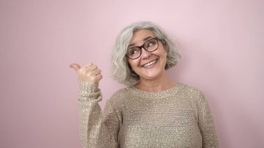 Middle age woman with grey hair smiling confident pointing to the side over isolated pink background
