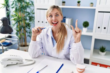 Beautiful dentist woman explaining how to brush teeth with toothbrush smiling with an idea or question pointing finger with happy face, number one 