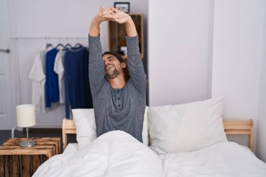 Young hispanic man waking up stretching arms at bedroom