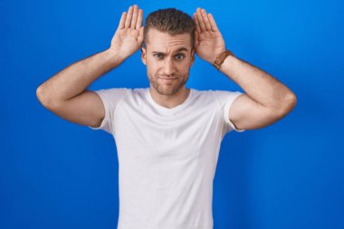Young caucasian man standing over blue background doing bunny ears gesture with hands palms looking cynical and skeptical. easter rabbit concept. 