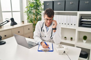 Young hispanic man doctor talking on smartphone writing medical report at clinic
