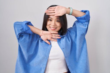 Beyaz arka planda duran genç Çinli bir kadın. Gülümseyen, güler yüzlü, yüzünü gösteren bir yuhalama oyunu oynayan. Şaşırdım ve çıktım. 