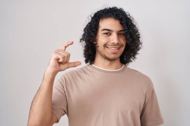 Beyaz arka planda duran kıvırcık saçlı İspanyol adam gülümsüyor ve el hareketleriyle küçük boy işareti yaparak kameraya bakıyor. ölçüm kavramı. 
