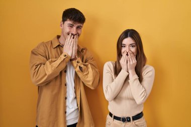 Sarı arka planda dikilen genç İspanyol çift gülüyor ve utanıyor kıkırdıyor elleriyle ağzını kapatıyor, dedikodu ve skandal konsepti yapıyor. 
