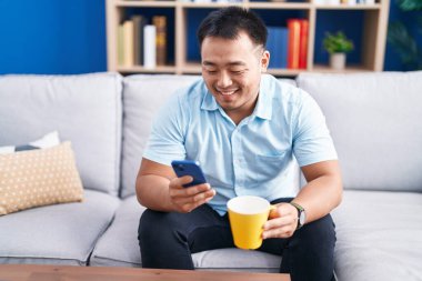 Genç Çinli adam akıllı telefon kullanıyor. Evde kahve içiyor.