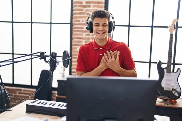 Young hispanic man playing piano keyboard at music studio smiling with hands on chest with closed eyes and grateful gesture on face. health concept.