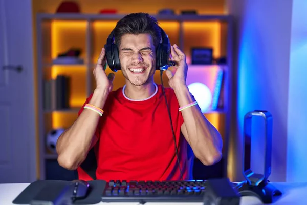 大音量の音楽のノイズのためにいらいらする表現で指で耳を覆うビデオゲームをしている若いヒスパニック系の男 聴覚障害の概念 — ストック写真