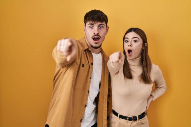Sarı arka planda dikilen genç İspanyol çift parmaklarını göstererek şaşırmış, ağzı açık, şaşırmış, ön tarafta bir şey var. 