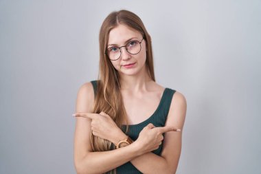 Beyaz arka planda duran beyaz tenli genç kadın her iki tarafı da parmaklarıyla işaret ediyor, farklı yönlere bakıyor. 