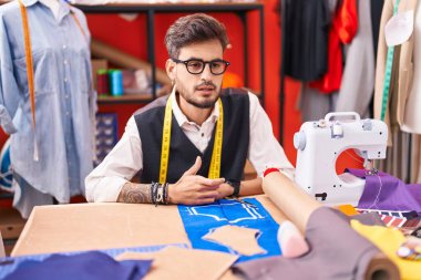 Young hispanic man tailor sitting on table speaking at atelier