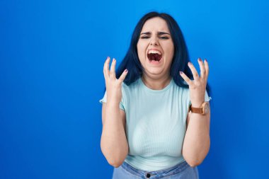 Young modern girl with blue hair standing over blue background crazy and mad shouting and yelling with aggressive expression and arms raised. frustration concept. 