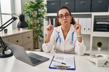 Doktor üniforması ve steteskop giyen genç İspanyol kadın üzgün ve üzgün görünüyor, parmaklarıyla yönü gösteriyor, mutsuz ve depresif.. 