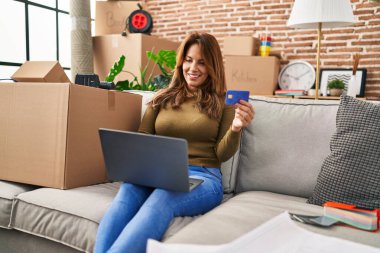 Laptop ve kredi kartı kullanan genç Latin kadın evdeki koltukta oturuyor.