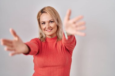 Sarışın bir kadın izole edilmiş arka planda durup kameraya bakarak sarılmak için kollarını açmış gülümsüyor. Mutluluğu kucaklayan neşeli bir ifade.. 