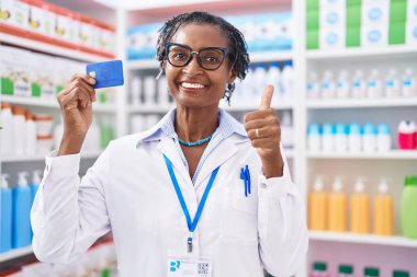 Eczanede çalışan Afrikalı kadın elinde kredi kartıyla mutlu ve pozitif gülümsüyor, başparmağıyla mükemmel ve onay işareti yapıyor. 
