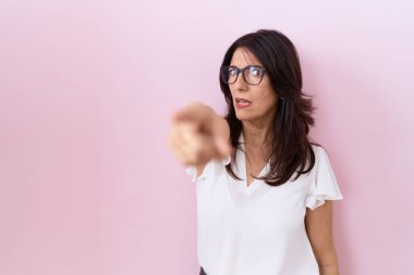 Orta yaşlı, beyaz tişörtlü ve gözlüklü bir İspanyol kadın kameraya kızgın ve sinirli bir şekilde bakıyor. 