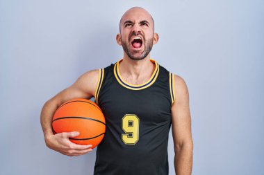 Young bald man with beard wearing basketball uniform holding ball angry and mad screaming frustrated and furious, shouting with anger. rage and aggressive concept. 