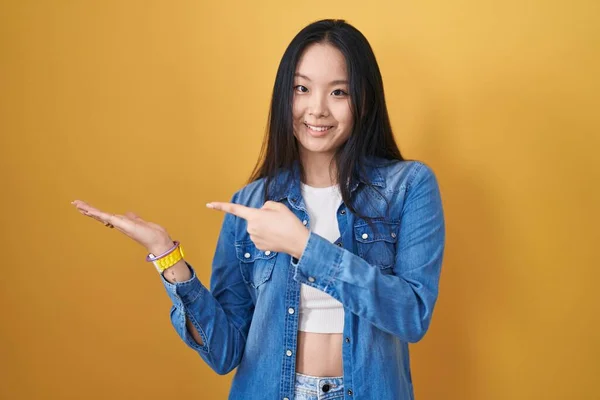 手で提示しながら 指で指している間に黄色の背景に立って若いアジアの女性は驚きとカメラに笑顔 — ストック写真