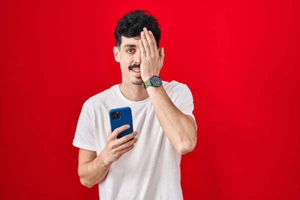 Hispanic Man Using Smartphone Red Background Covering One Eye Hand — 스톡 사진