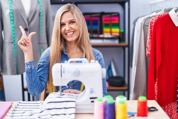 Blonde woman dressmaker designer using sew machine with a big smile on face, pointing with hand finger to the side looking at the camera.