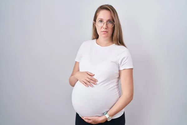 赤ん坊を期待して妊娠中の若い女性は 顔に深刻な表情でリラックスした妊娠中の腹に触れます カメラを見ているだけで — ストック写真