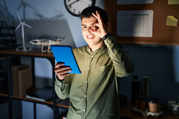 Non binary person using touchpad device at night doing ok gesture with hand smiling, eye looking through fingers with happy face.