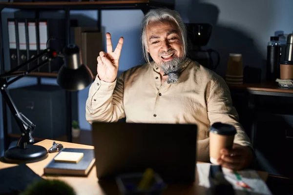 深夜オフィスで働く白髪の中年男性は 指で勝利サインをするカメラに顔をウィンクし幸せな笑顔で — ストック写真
