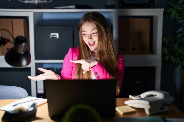 Geceleri ofiste çalışan beyaz bir kadın hayret içinde kameraya bakıp gülümserken eliyle ve parmağıyla işaret ediyor.. 