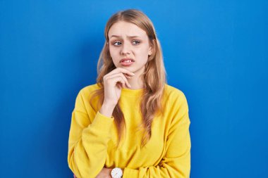 Young caucasian woman standing over blue background thinking worried about a question, concerned and nervous with hand on chin 