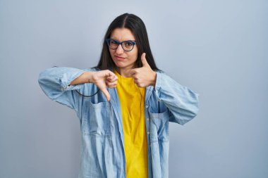 Latin kökenli genç bir kadın mavi arka planda dikiliyor. Baş parmaklarını yukarı aşağı hareket ettiriyor. Anlaşmazlık ve uzlaşma ifadesi. çılgın çatışma 