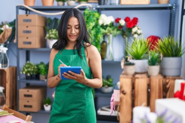 Young hispanic woman florist smiling confident using touchpad at florist shop